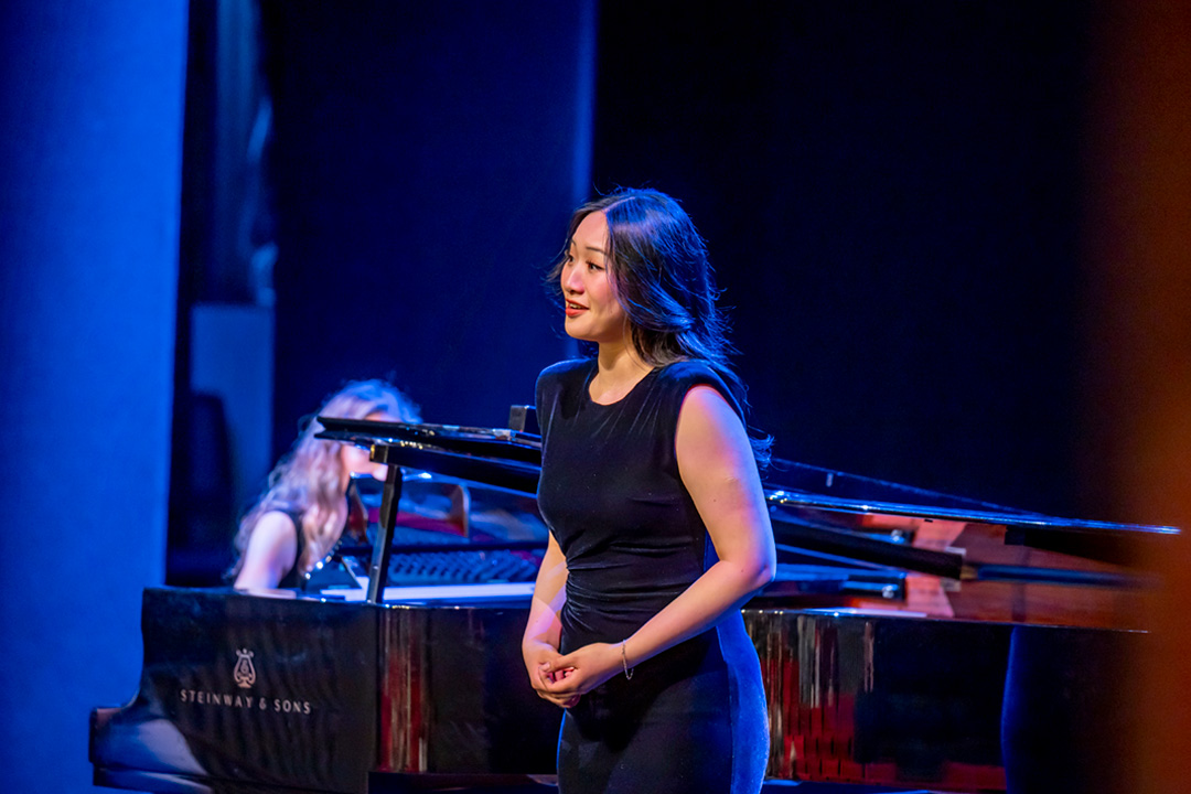 A female student wearing a nice dress, singing on stage, with a piano accompanying her.